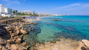 A beach with clear turquoise water, rocky shoreline, and a row of hotel buildings in the background. - Sunset Villa in Protaras (Paralimni)