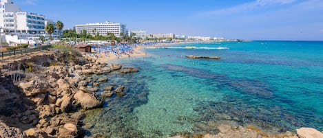 A beach with clear turquoise water, rocky shoreline, and a row of hotel buildings in the background.