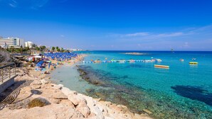 A beach with clear turquoise water, blue umbrellas, and boats. - Sunset Villa in Protaras (Paralimni)