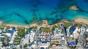 An aerial view of a coastal resort with multiple buildings, a beach, and clear blue waters. - Silver Sands Penthouse (Paralimni)