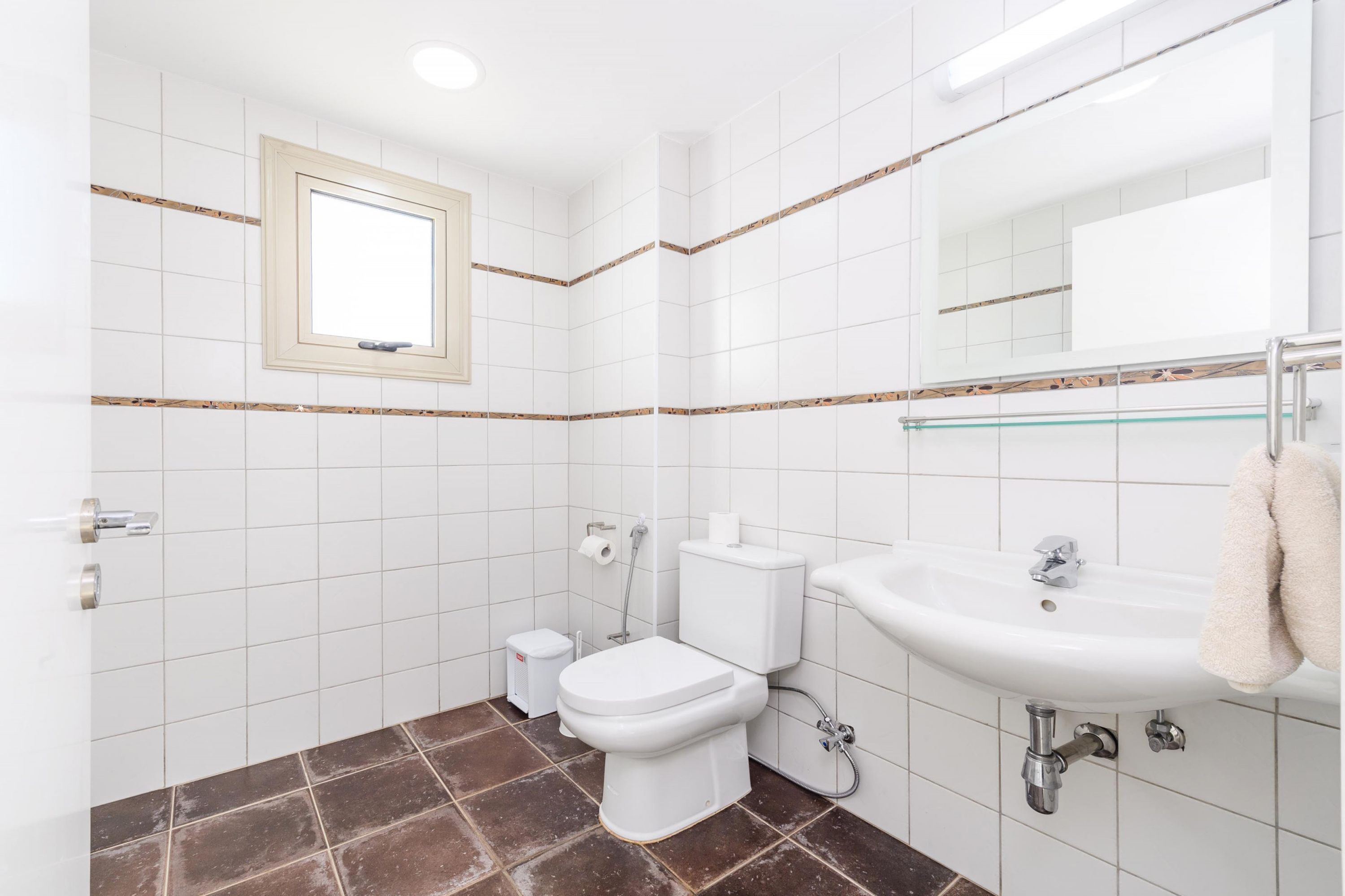 Ein Badezimmer mit weißen Fliesen, einer Toilette, einem Waschbecken und einem Fenster.