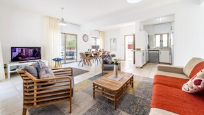 A modern living room with a TV, wooden furniture, a dining area, and a kitchen. - Sunrise Bay Villa 1 (Paralimni)