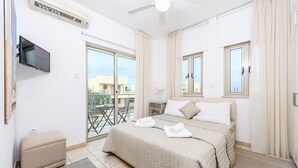 A hotel room with a bed, a flat-screen TV, a small sofa, a balcony with a table and chairs, and a ceiling fan. - Sunrise Bay Villa 6 (Paralimni)