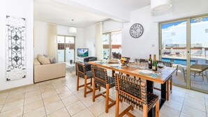 A modern living room with a sofa, TV, dining table, chairs, and a view of the pool. - Sunrise Bay Villa 6 (Paralimni)