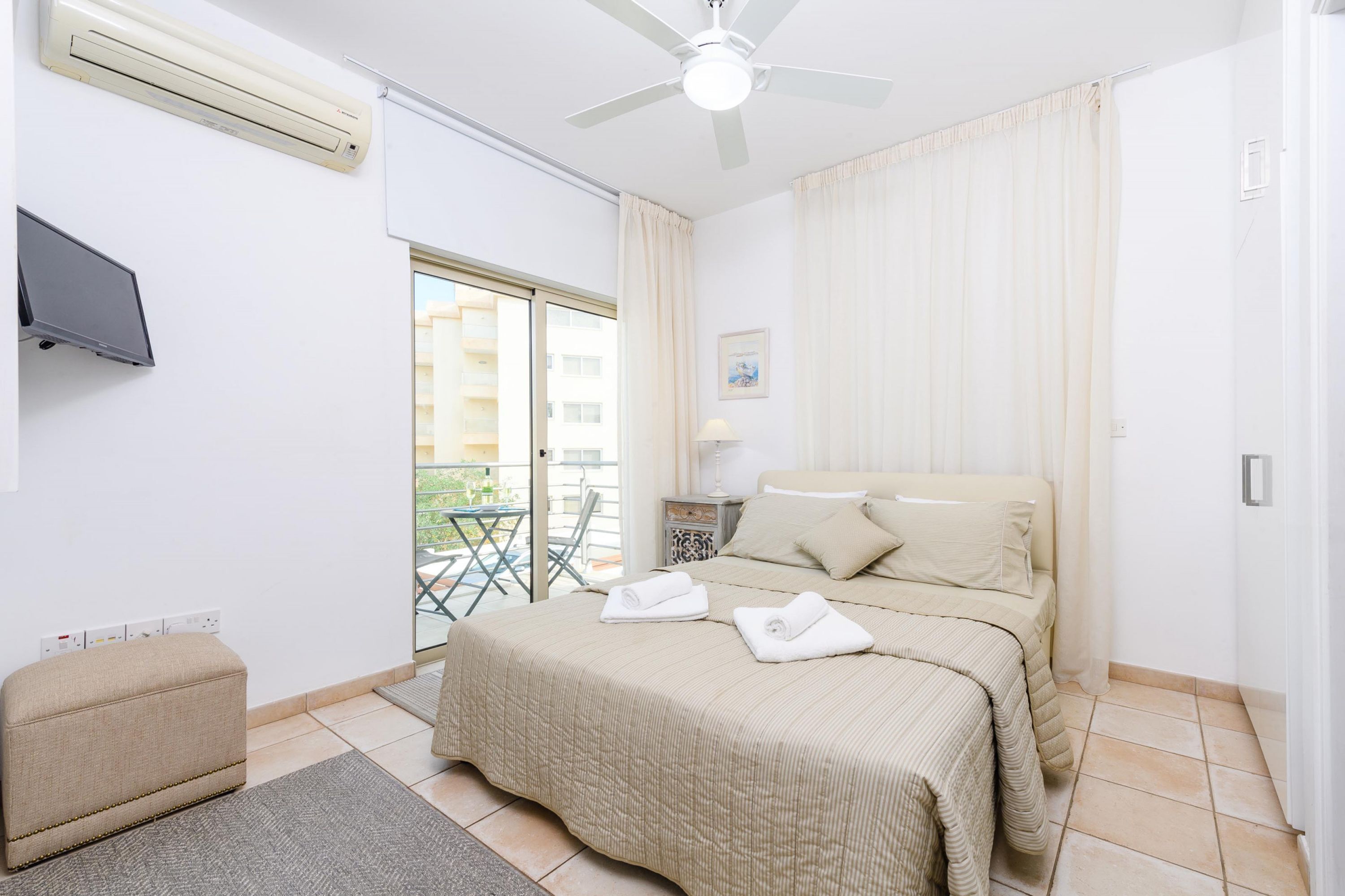 Ein Hotelzimmer mit Bett, Fernseher, Klimaanlage, Deckenventilator, Balkon und Blick auf Gebäude.