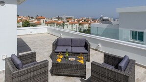Terraza en la azotea con mobiliario de mimbre, una mesa con una botella y vasos, y vistas de edificios al fondo.