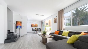 Uma sala de estar moderna com um sofá cinza, uma mesa de centro e um espaço de jantar com uma mesa e cadeiras. Há uma escada à esquerda e uma janela grande à direita.