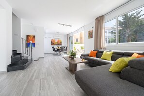 Una sala de estar moderna con un sofá gris, una mesa de centro y un comedor con una mesa y sillas. Hay una escalera a la izquierda y un ventanal grande a la derecha.