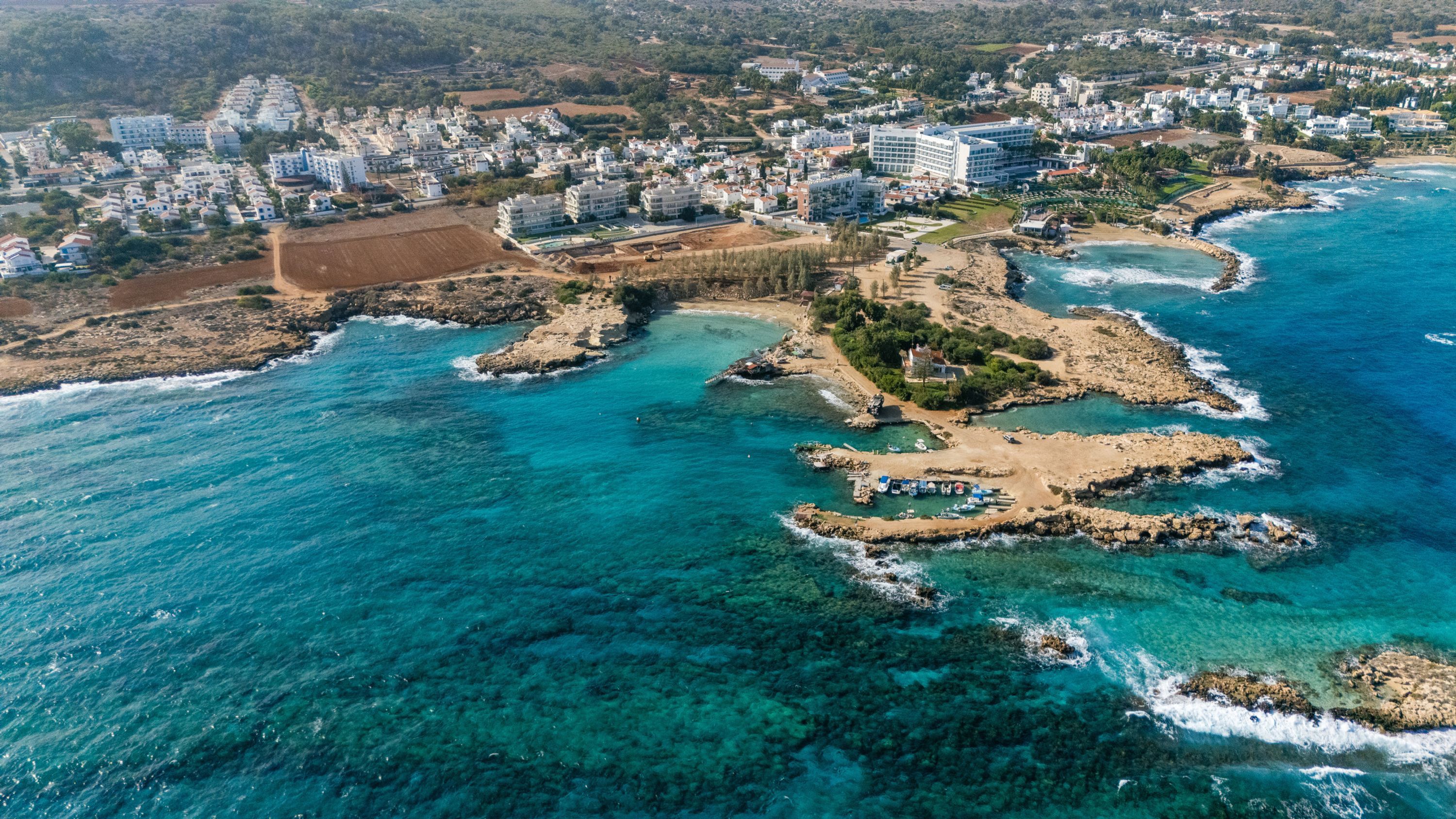 An aerial view of a coastal town with clear blue waters, rocky outcrops, and a mix of residential and commercial buildings.