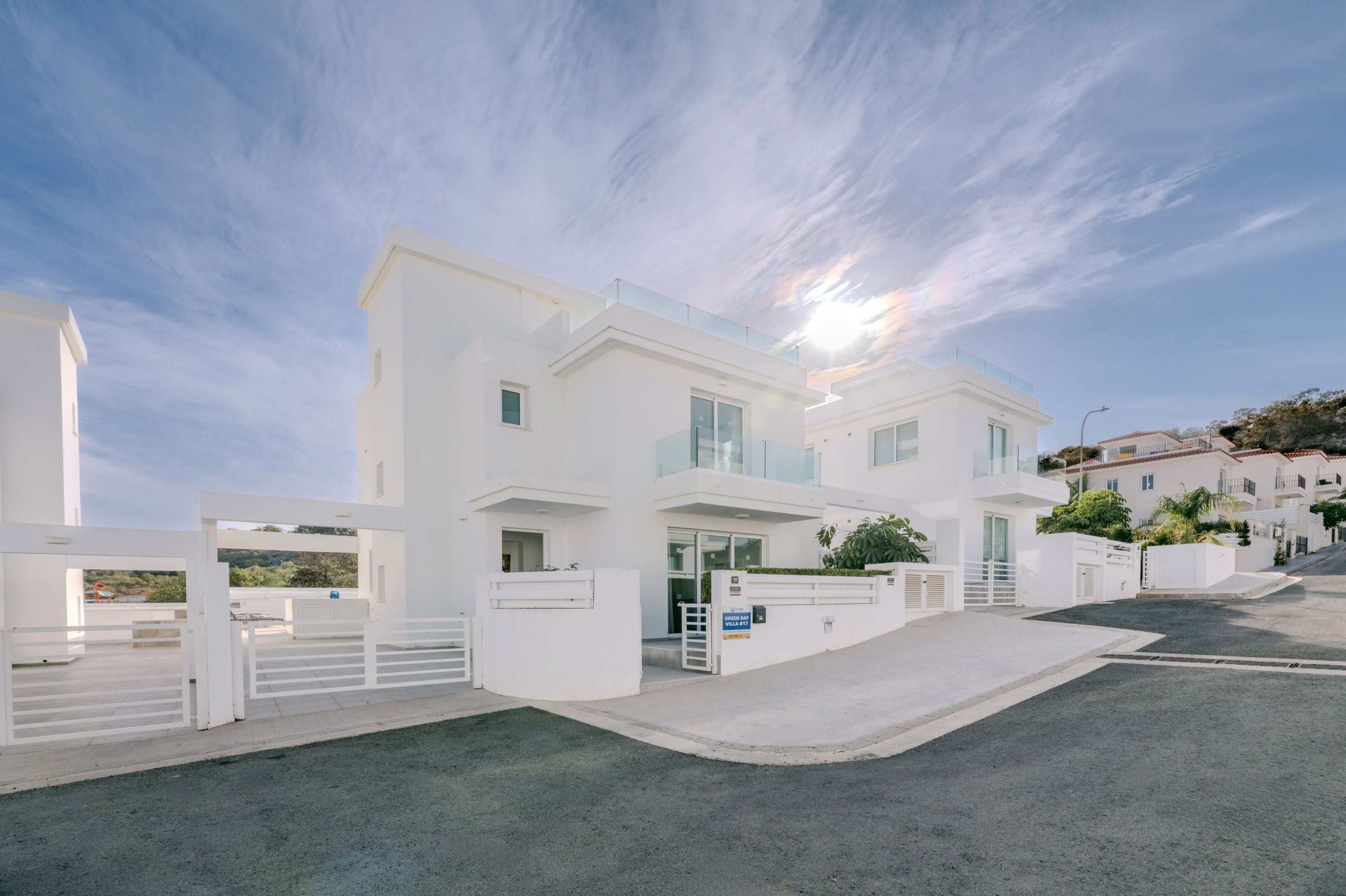 A modern white villa with a glass balcony, a driveway, and a clear sky.