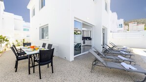 A modern outdoor dining area with a table and chairs, a kitchen visible through an open door, and a row of sun loungers with towels. - Green Bay Villa 17 in Protaras (Paralimni)
