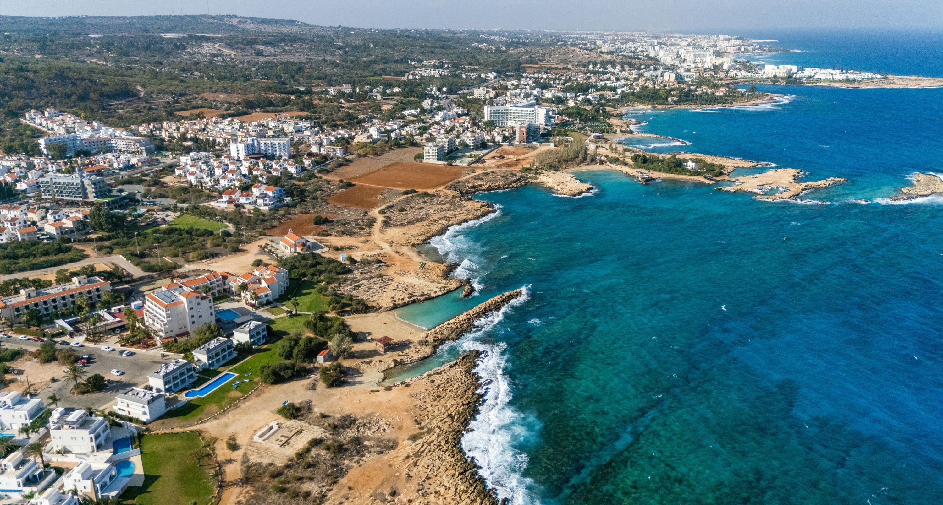 An aerial view of a coastal town with residential buildings, a beach, and clear blue waters.