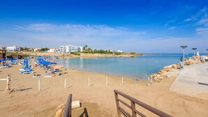 A beach with sun loungers and umbrellas, a rocky shoreline, and buildings in the background. - Villa Levanda 40 in Protaras (Paralimni)
