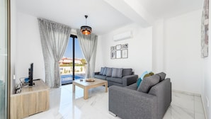 A modern living room with a grey sofa, a wooden coffee table, and a TV on a wooden stand. There is a large window with curtains, an air conditioning unit, and framed pictures on the wall. - Villa Levanda 40 in Protaras (Paralimni)