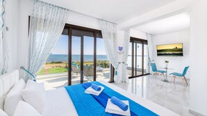 A hotel room with a large bed, a balcony with a view of the sea, and a dining area with two chairs. - Serenity Seafront Villa (Sotira)