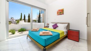 Un dormitorio moderno con un amplio ventanal corredera que da a un balcón, una cama con colcha colorida, una mesita de noche y una obra de arte colgada en la pared.