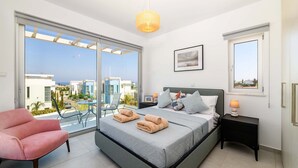 A modern bedroom with a large bed, a pink chair, and a balcony with a view of buildings. - Mylos Lifestyle Seaview Villa 21 (Paralimni)