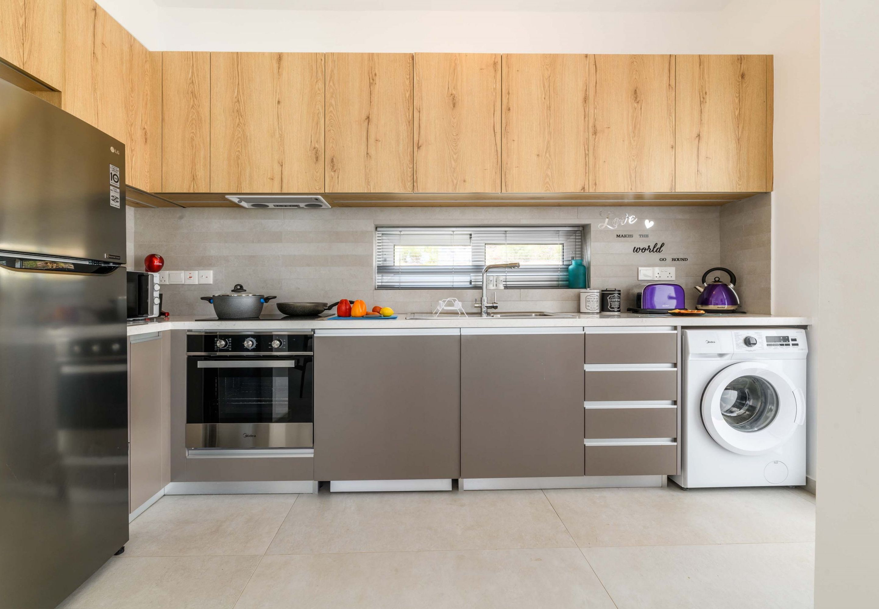 A modern kitchen with wooden cabinets, a built-in oven, a washing machine, and a window with blinds.