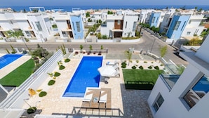 A modern residential area with a central swimming pool, surrounded by well-maintained lawns and a few trees. - Mylos Lifestyle Seaview Villa 21 (Paralimni)