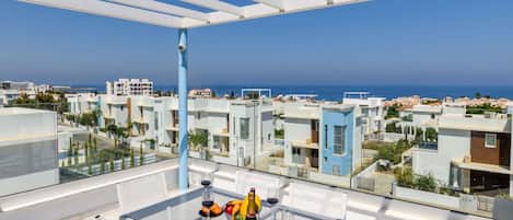 A modern outdoor terrace with a glass table and white chairs, overlooking a coastal cityscape.