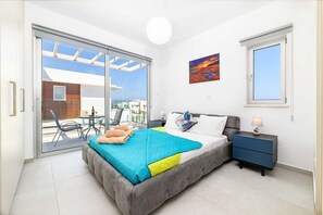 A bedroom with a bed, a nightstand, a window, and a sliding glass door leading to an outdoor area. - Mylos Lifestyle Seaview Villa 21 (Paralimni)