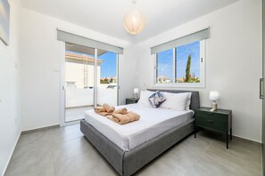 A modern bedroom with a large bed, a nightstand, a lamp, and a view of the outside. - Mythical Sands Ae01 in Protaras (Paralimni)