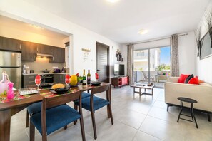 A modern kitchen with dark wooden cabinets, a dining area with blue chairs, and a living room with a beige sofa and red cushions. - Mythical Sands Ae01 in Protaras (Paralimni)