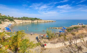 A beach with clear blue water, people swimming and sunbathing, and a rocky coastline. - Mythical Sands Ae01 in Protaras (Paralimni)