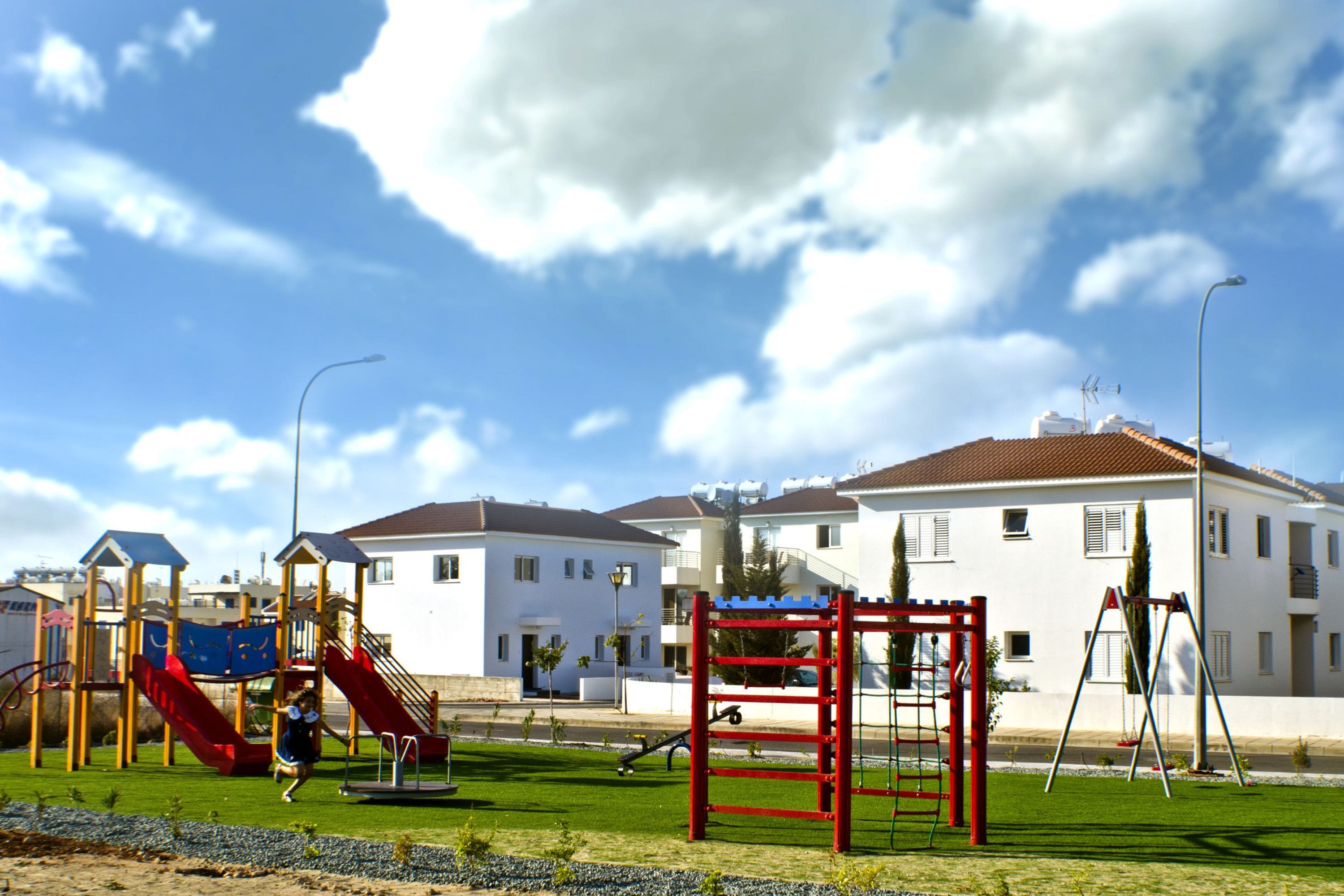 A playground with slides and climbing structures, surrounded by a grassy area and a row of white buildings with red roofs.