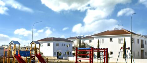 A playground with slides and climbing structures, surrounded by a grassy area and a row of white buildings with red roofs.