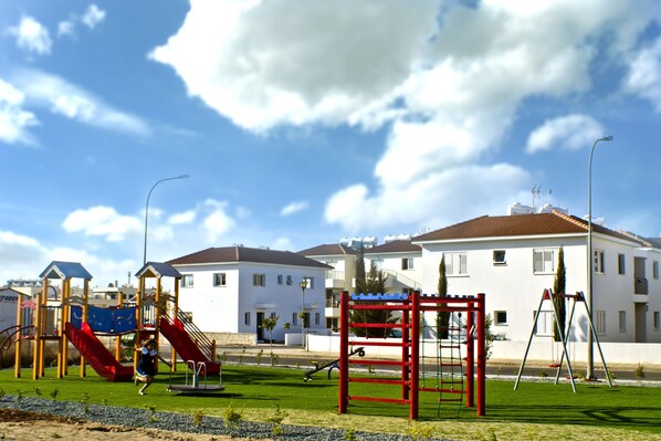 A playground with slides and climbing structures, surrounded by a grassy area and a row of white buildings with red roofs. - Mythical Sands Ae01 in Protaras (Paralimni)