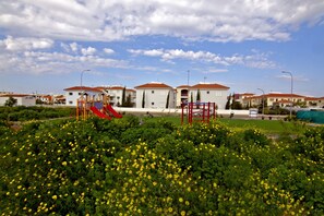 A residential area with apartment buildings, a playground, and a garden with yellow flowers. - Mythical Sands Ae01 in Protaras (Paralimni)