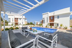 Un área de piscina moderna al aire libre con pérgolas blancas, mobiliario exterior gris y un cielo azul despejado.