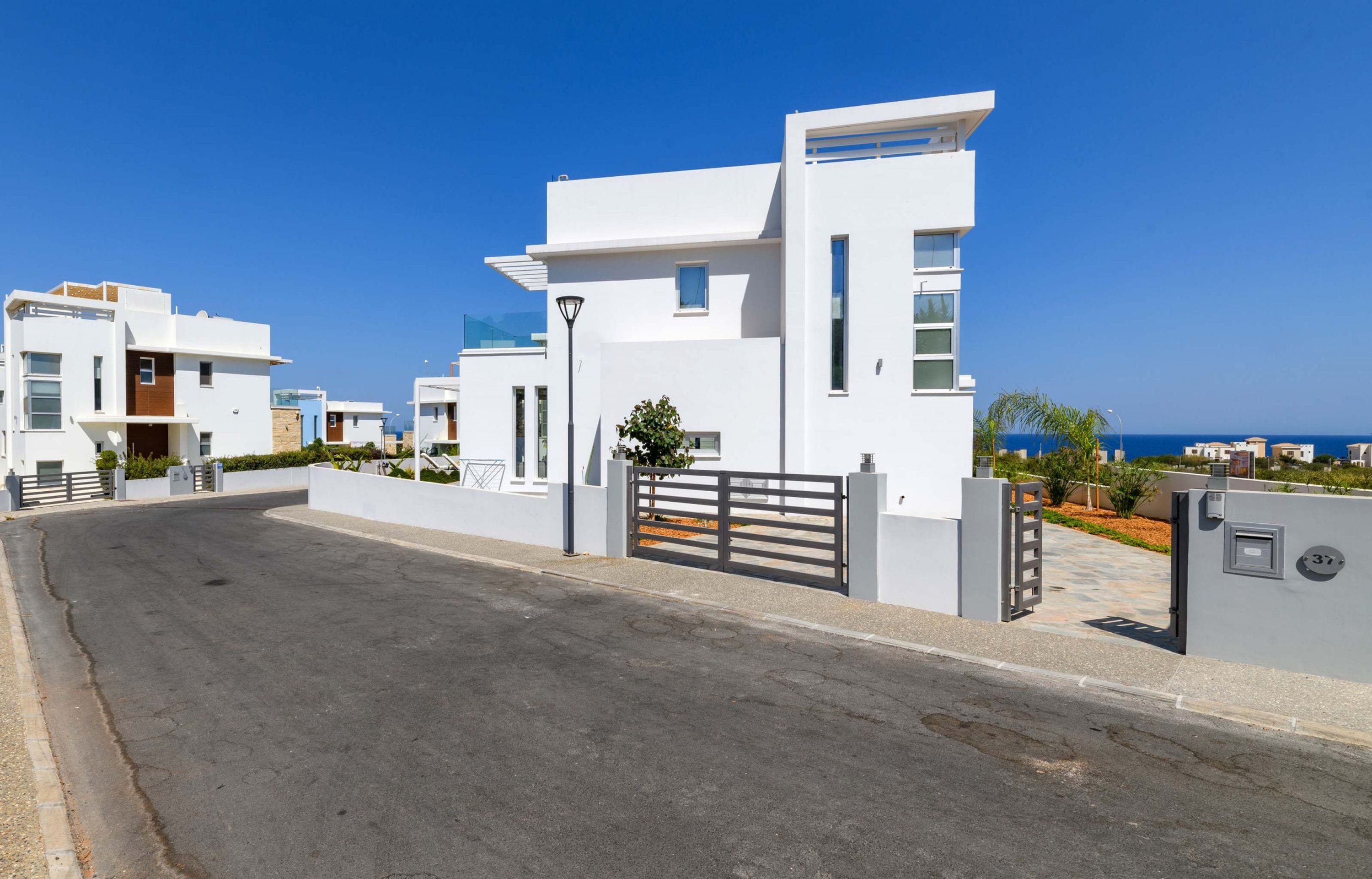 A modern residential building with a gated entrance, a clear view of the street, and a coastal backdrop.