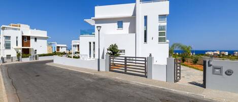 A modern residential building with a gated entrance, a clear view of the street, and a coastal backdrop.