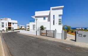 Un edificio residencial moderno con entrada porticada, vista despejada a la calle y un paisaje costero de fondo.