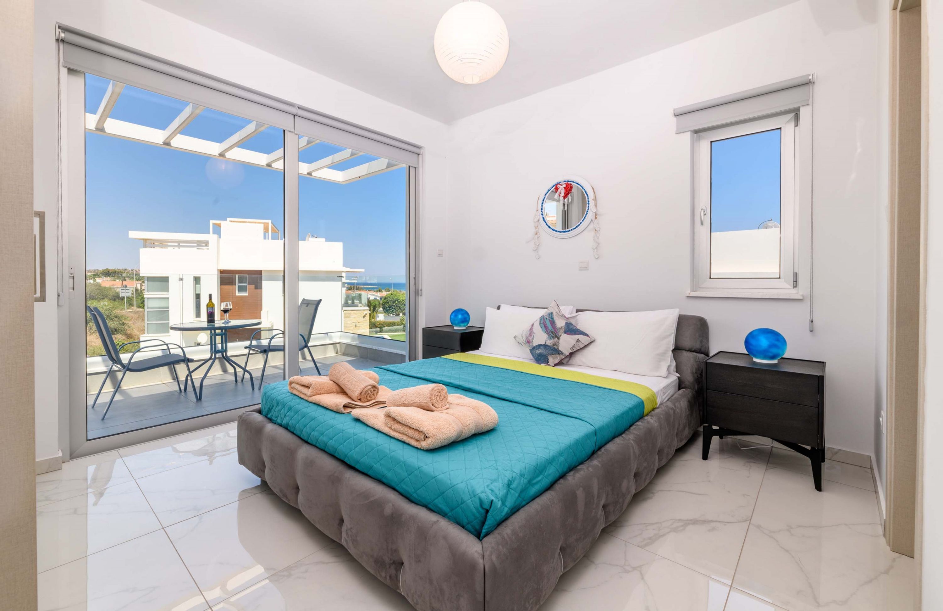 A bedroom with a bed, a bedside table, a nightstand, a mirror, and a view of the outside.
