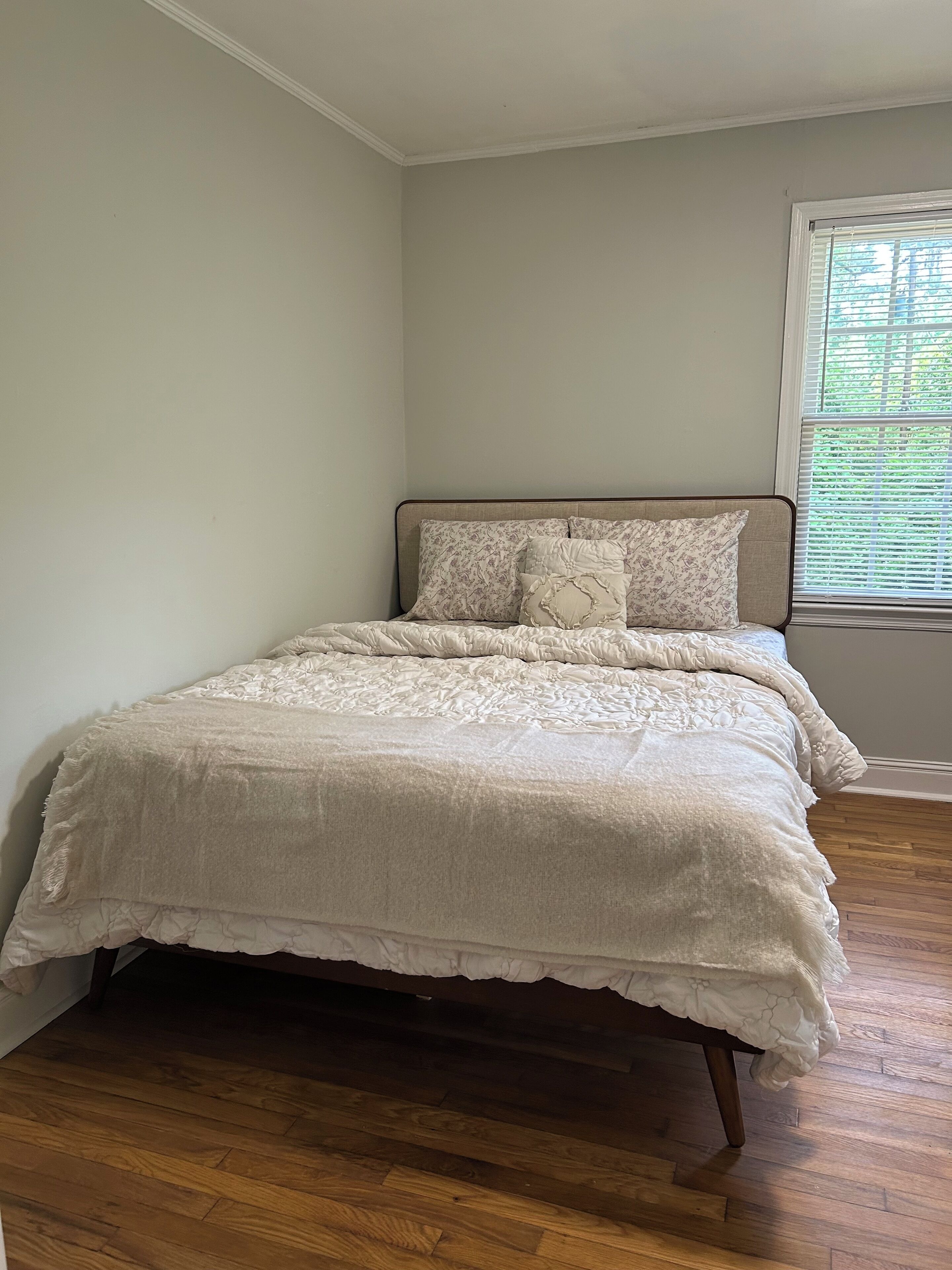 Un dormitorio con suelo de madera, una cama con colcha blanca y dos almohadas, una ventana con persianas y una pared lisa.