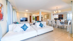 A modern living room with a white sofa set, a flat-screen TV, and a dining area with a chandelier. - Royal Beachfront Villa (Paralimni)