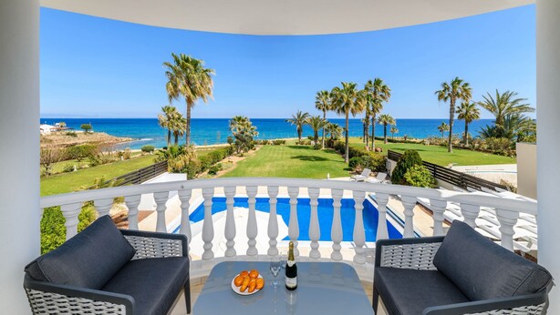 A balcony with a pool, two chairs, a table with a bottle and oranges, and a view of the ocean and palm trees. - Royal Beachfront Villa (Paralimni)