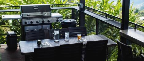 An outdoor dining area with a grill, table, chairs, and a view of lush greenery.