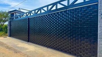 A modern metal fence with a geometric pattern, a concrete pathway, and a building with a stone facade in the background.