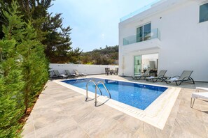 A modern outdoor swimming pool with a clear blue water feature, surrounded by lounge chairs and a white building with a glass balcony. - Green Bay Villa 16 in Protaras (Paralimni)