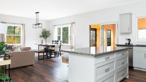 A modern kitchen with white cabinetry, a granite countertop, and a dining area with a table and chairs. - All Season Muskoka Cottage*HotTub*Sauna*Beach*BBQ (Huntsville)