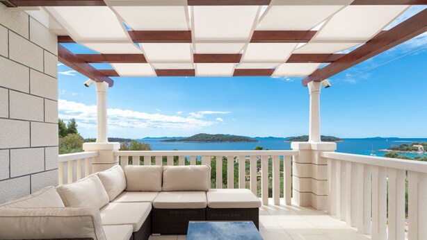 A covered outdoor seating area with a sofa and a table, overlooking a scenic view of a body of water and distant land.