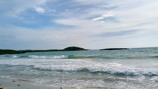 Una playa con olas, una orilla arenosa y unas islas a lo lejos.