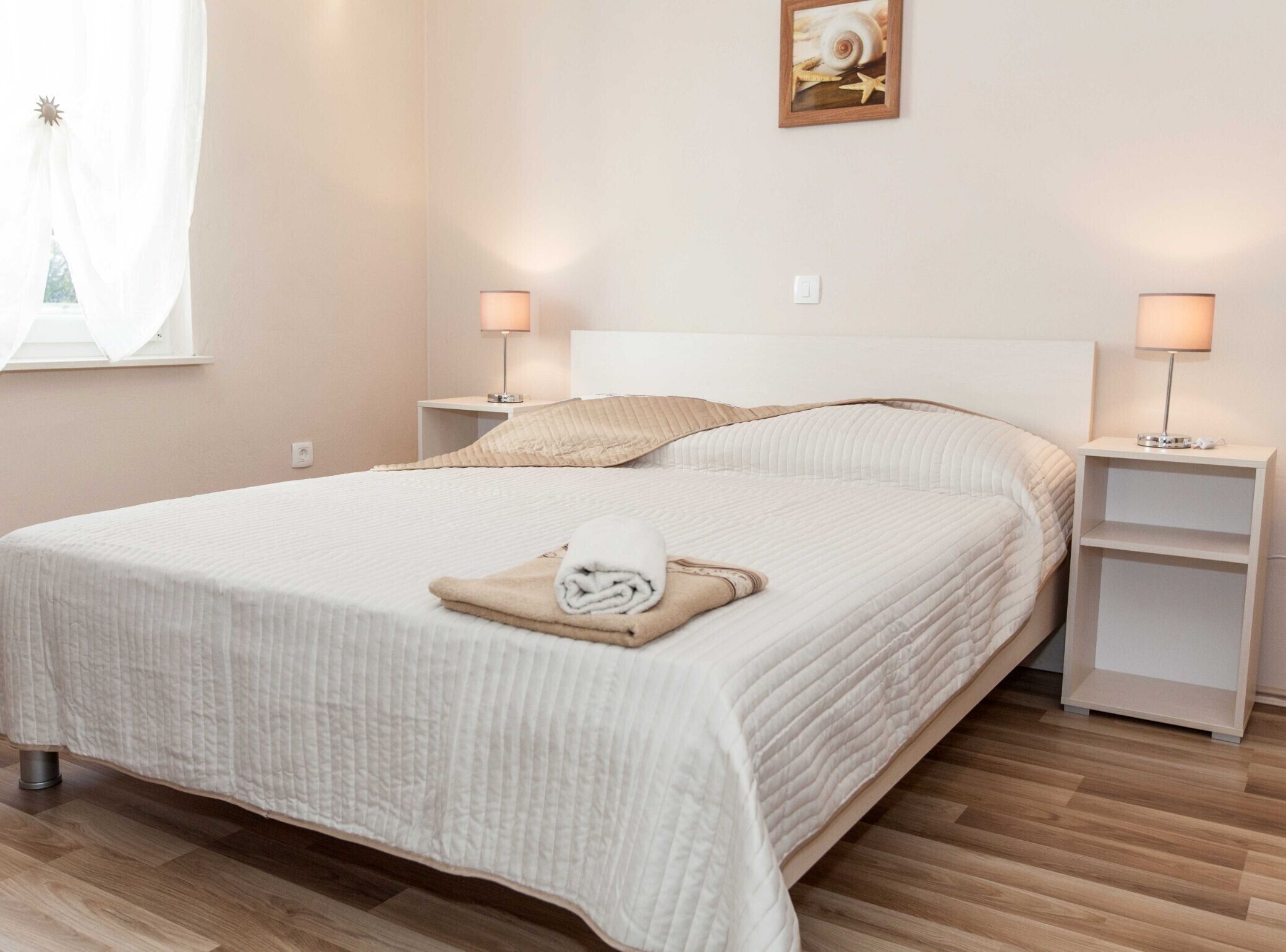 A neatly made bed with a folded towel, two bedside tables with lamps, a framed picture on the wall, and a window with sheer curtains.