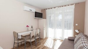 Una sala de estar moderna con un sofá, una mesa de comedor con sillas, un televisor de pantalla plana montado en la pared y una ventana con cortinas translúcidas.