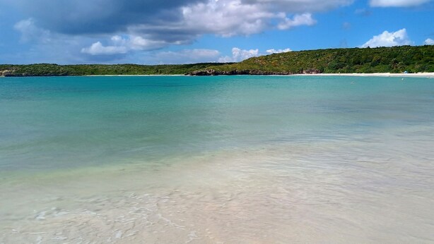 A beach with clear turquoise water, a sandy shore, and a forested hill in the background.