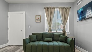 A living room with a green sofa, a framed picture on the wall, and a window with curtains. - Serenity Stays Charming 3bd 2.5ba Oasis (Philadelphia)